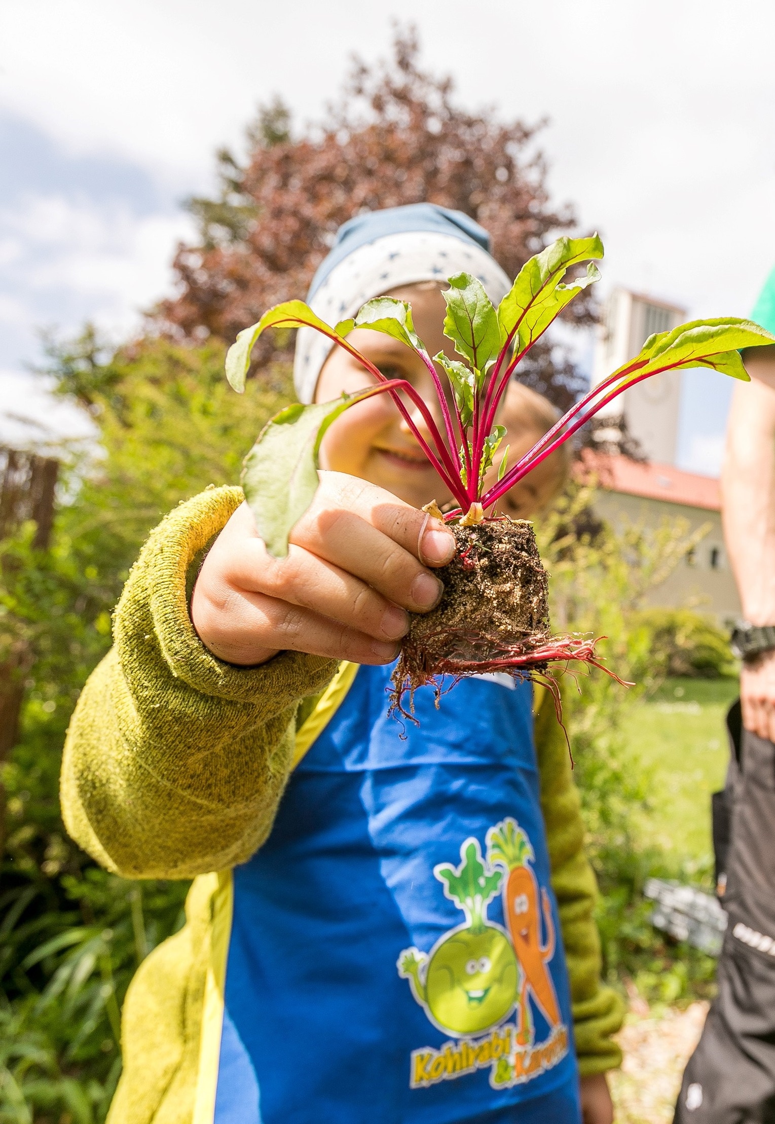 Kita-Kind zeigt einen Setzling in die Kamera bei "Gemüsebeete für Kids" der EDEKA Stiftung.