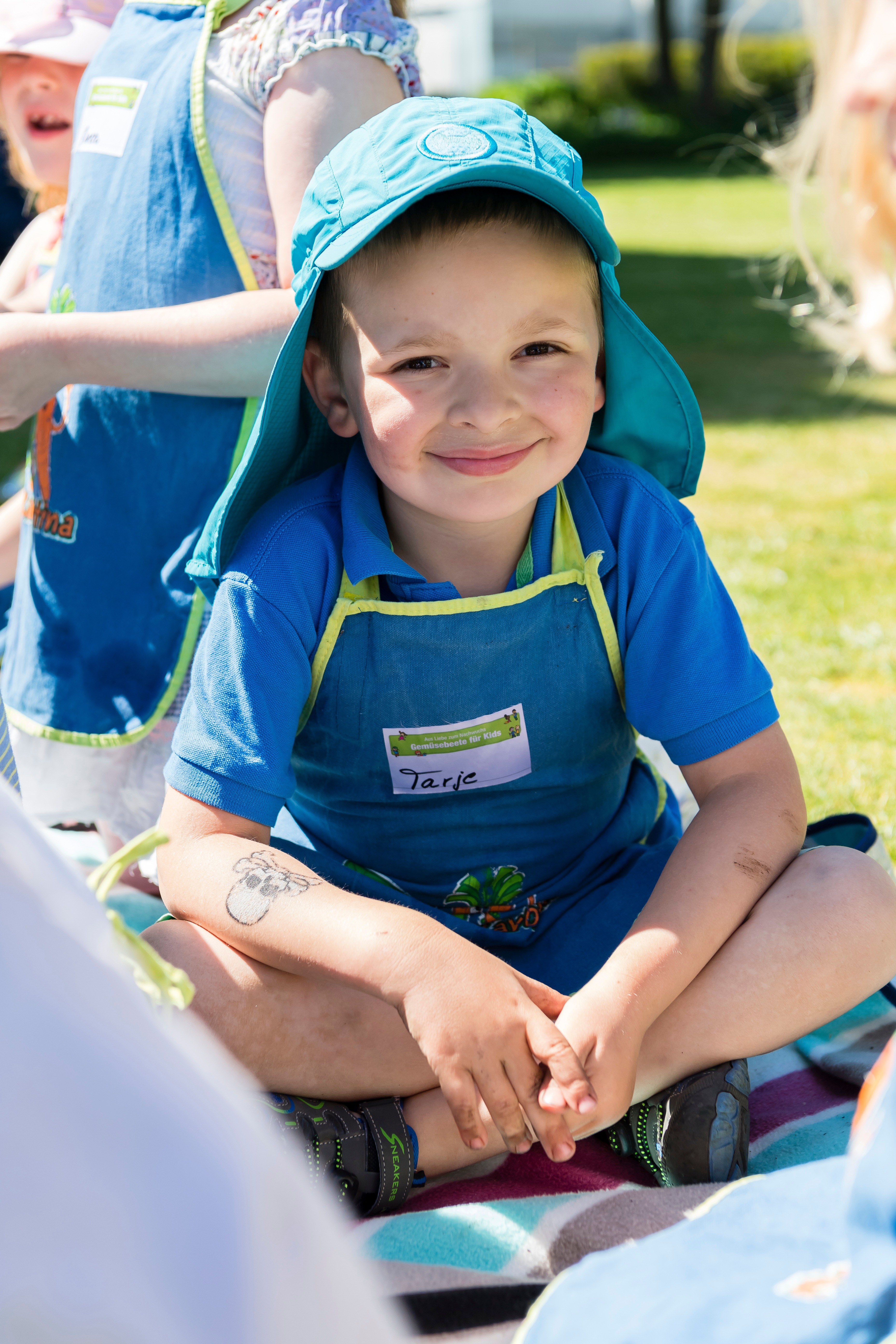 Ein Kind in einer blau-gelben Schürze sitz auf dem Rasen und lächelt bei "Gemüsebeete für Kids".