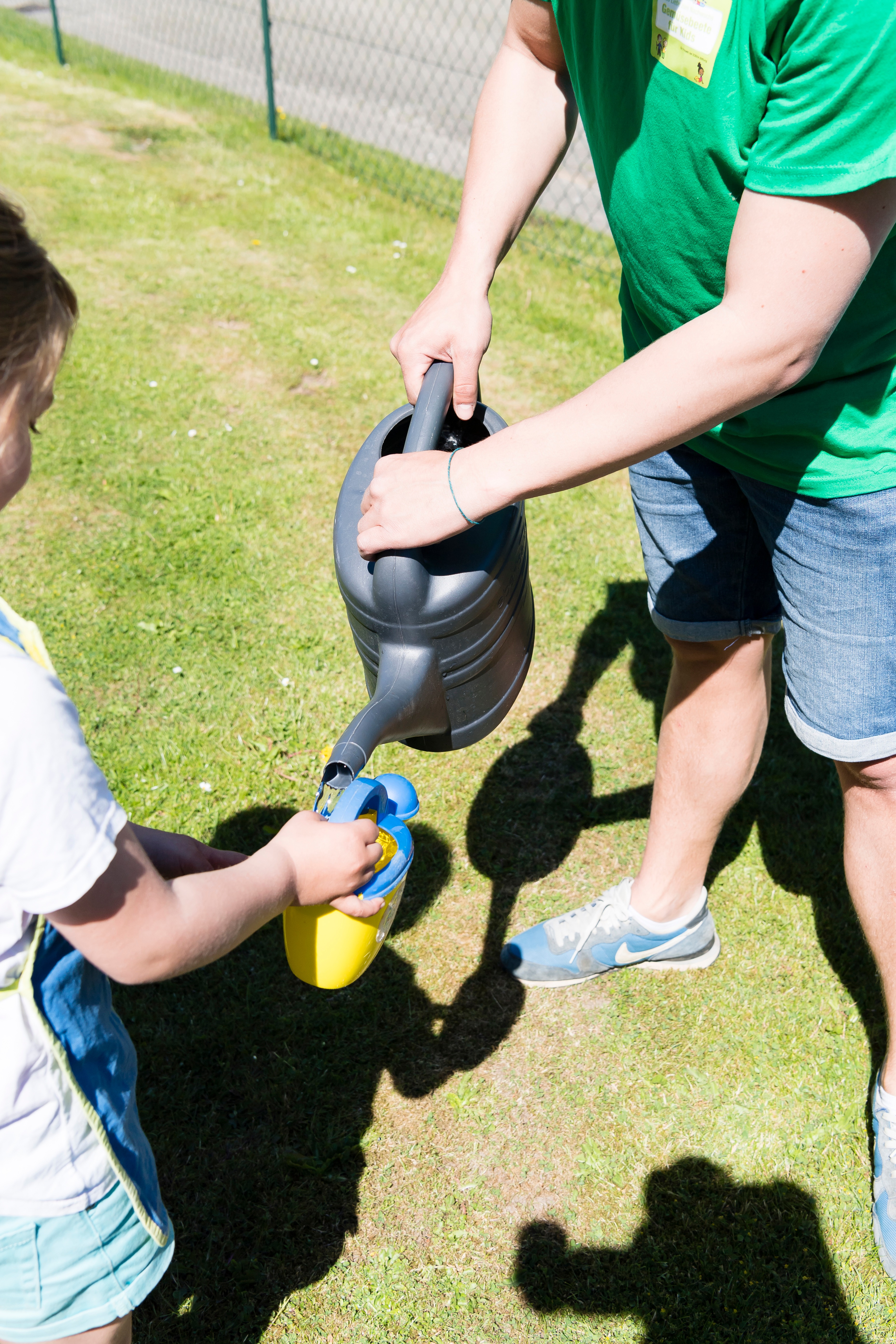 Eine kleine blau-gelbe Kinder-Gießkanne wird aus einer großen Gießkanne aufgefüllt bei "Gemüsebeete für Kids".