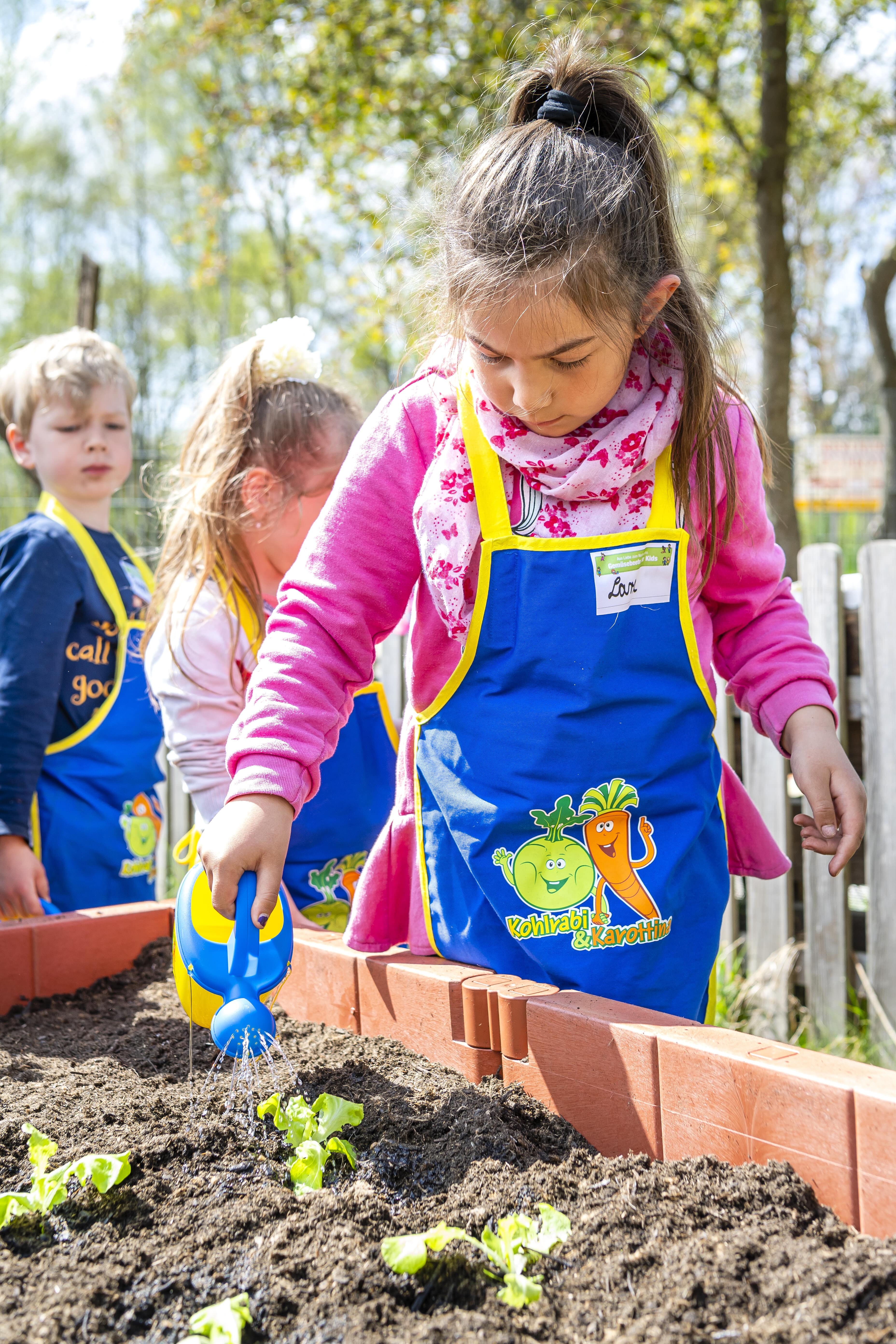 Kita-Kinder stehen um ein Hochbeet herum, ein Mädchen gießt die frisch gepflanzten Setzlinge bei "Gemüsebeete für Kids".